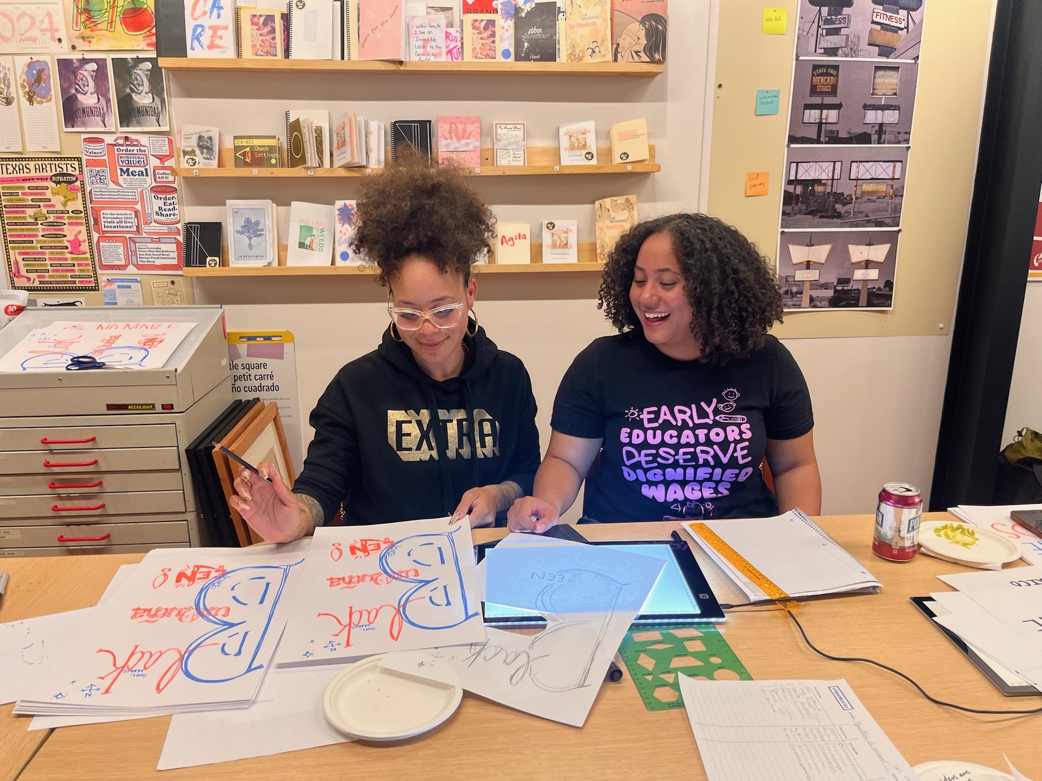 Two people sit side by side at a table filled with art supplies and works in progress.