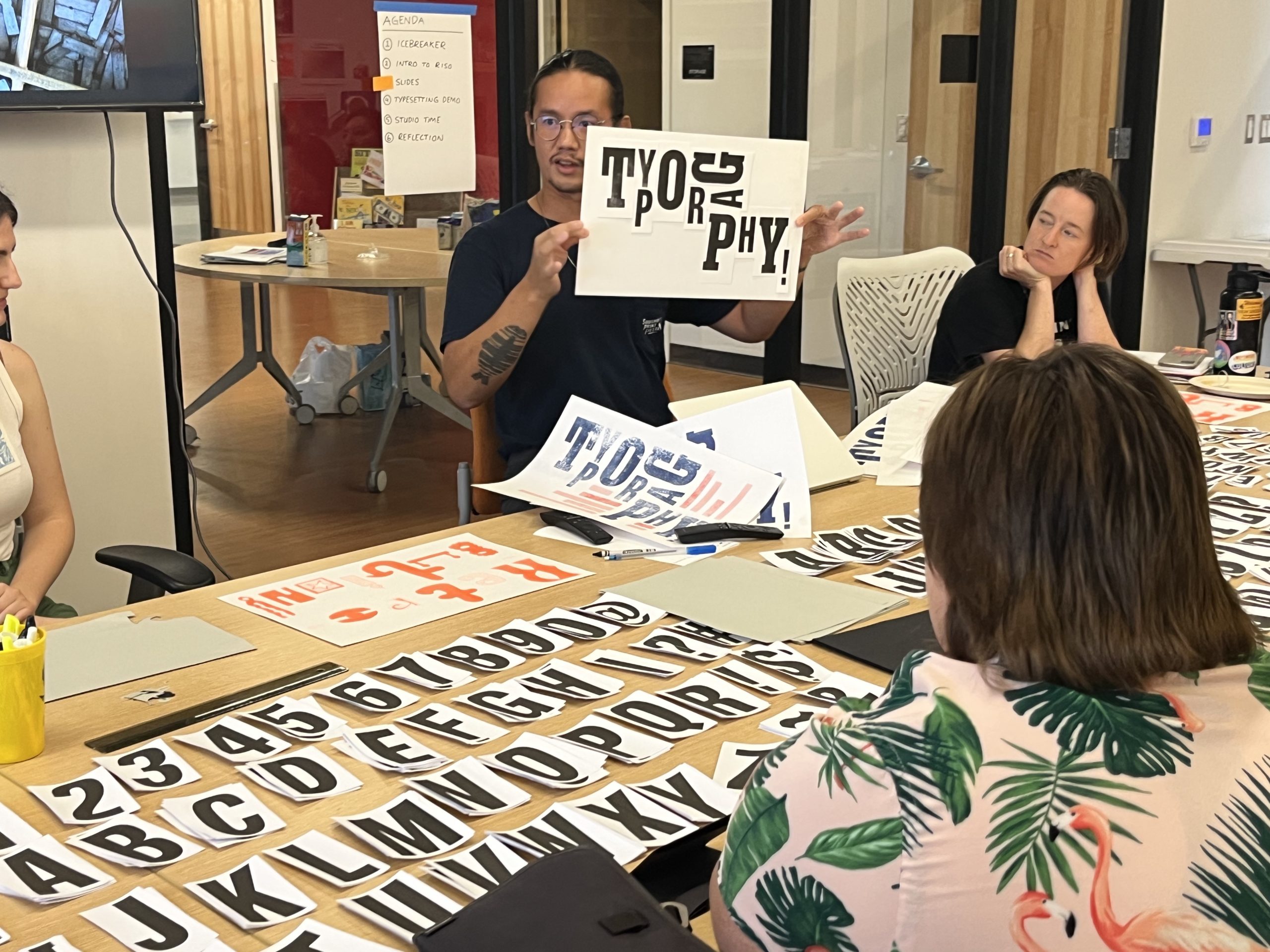 A person holds up a sign that says "typography" in a variety of fonts, as workshop participants look on.