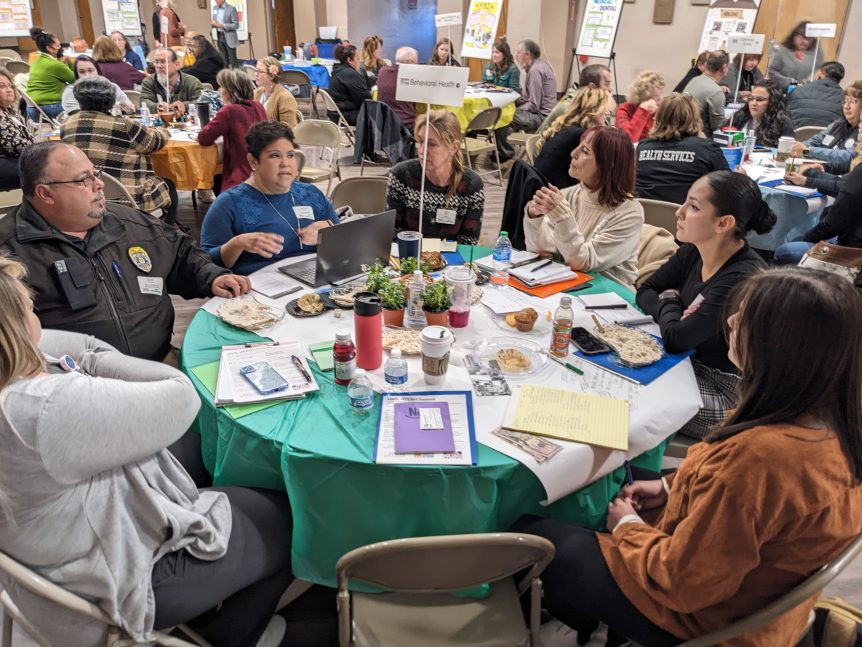 Foreground: diverse group of adults sitting at a roundtable dedicated to the topic of behavioral health, engaged in conversation. Background: a room full of similar tables, with adults engaged in focused conversations. Food, beverages, and notetaking materials are on the tables, and posters and flip-charts line the boundaries of the room.