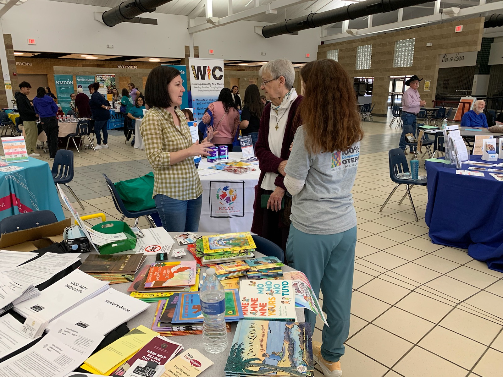 Foreground: Three women standing and talking near a table of early childhood reading materials. Background: posters for public resources for families and children, similar small groups of adults standing and talking at various tables filled with educational materials.