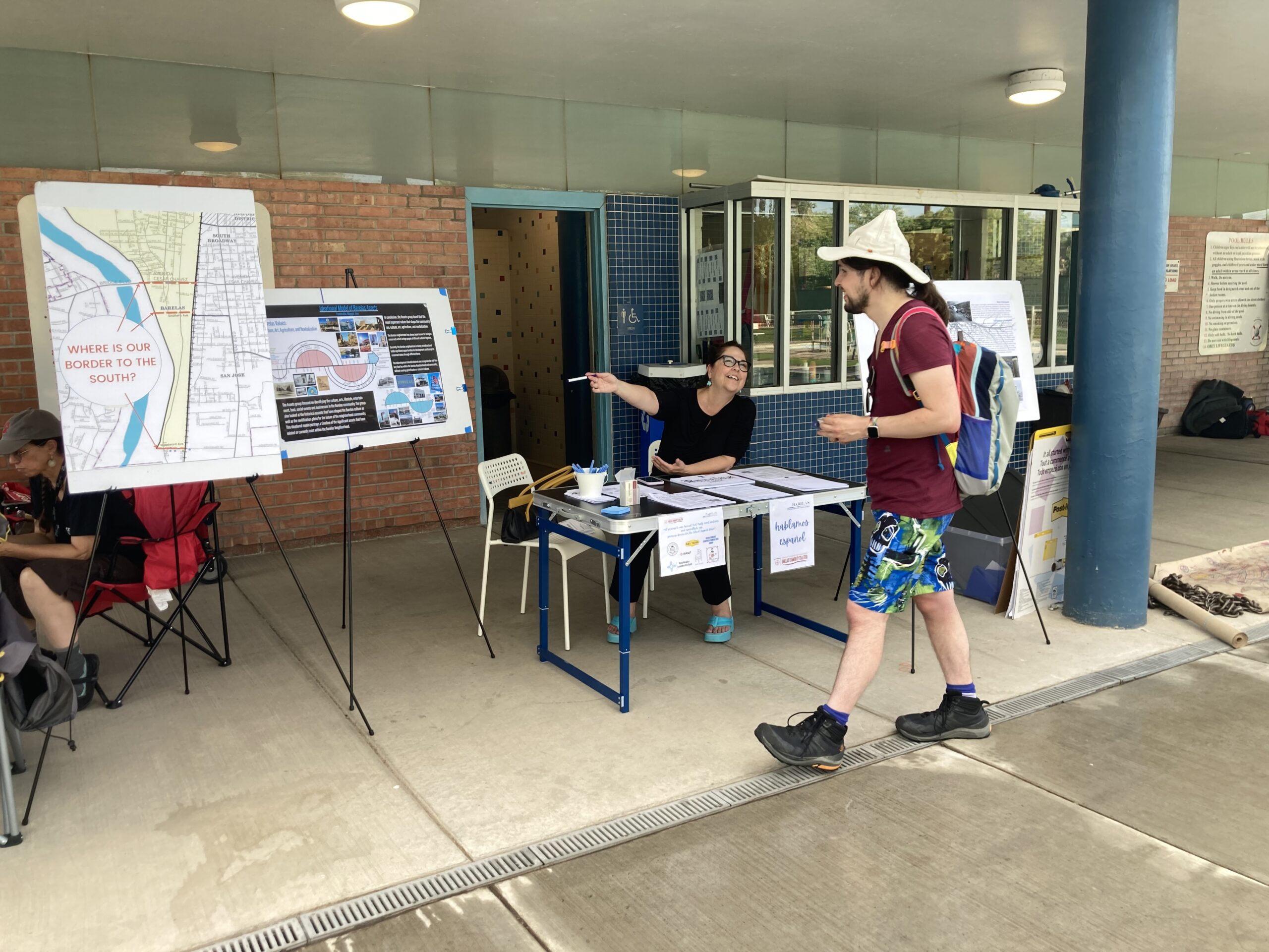 A person arrives at the Barelas Neighborhood Association pool party, and is welcomed by another person seated at a table, gesturing toward a neighborhood map and a poster with additional information about the community.